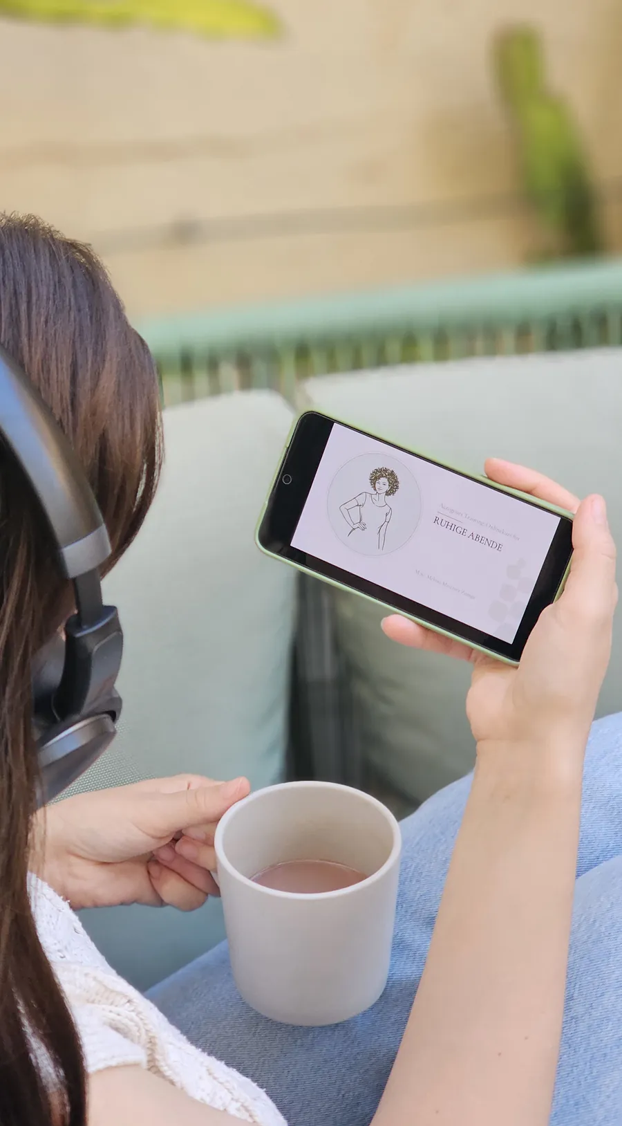 Frau mit Kopfhoerern, Tasse in der einen und Smartphone in der anderen Hand